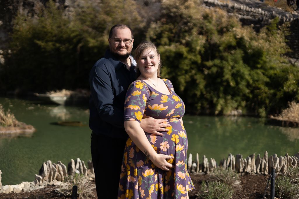Couple posing outdoors by a green river, woman in floral dress cradling baby bump, man in navy shirt embracing her.