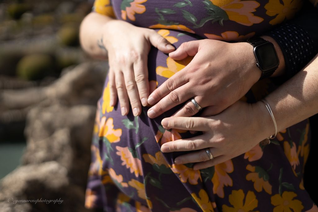 Couple tenderly cradling pregnant belly in floral maternity dress outdoors, showcasing wedding rings and bracelets.