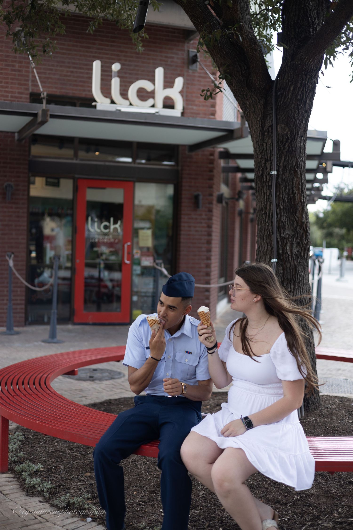People in white and blue outfits sitting on red curved benches outside a brick storefront.