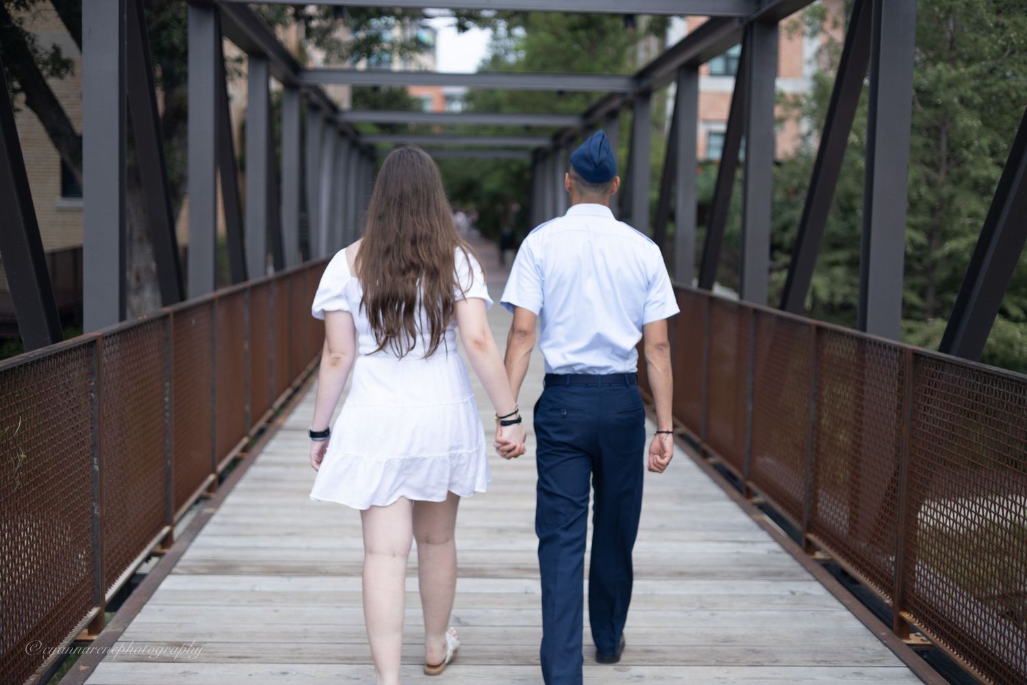 Couple holding hands while walking across an outdoor metal bridge covered in rust and foliage.
