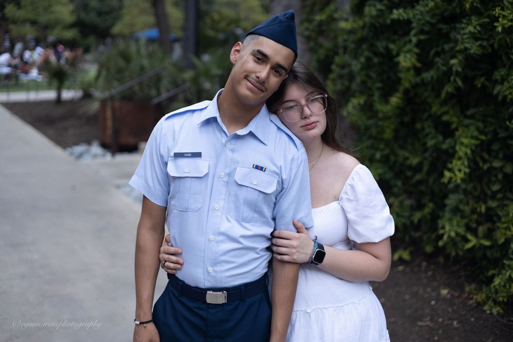 A military service member in dress blues stands with a companion in a white dress on a paved path near greenery.
