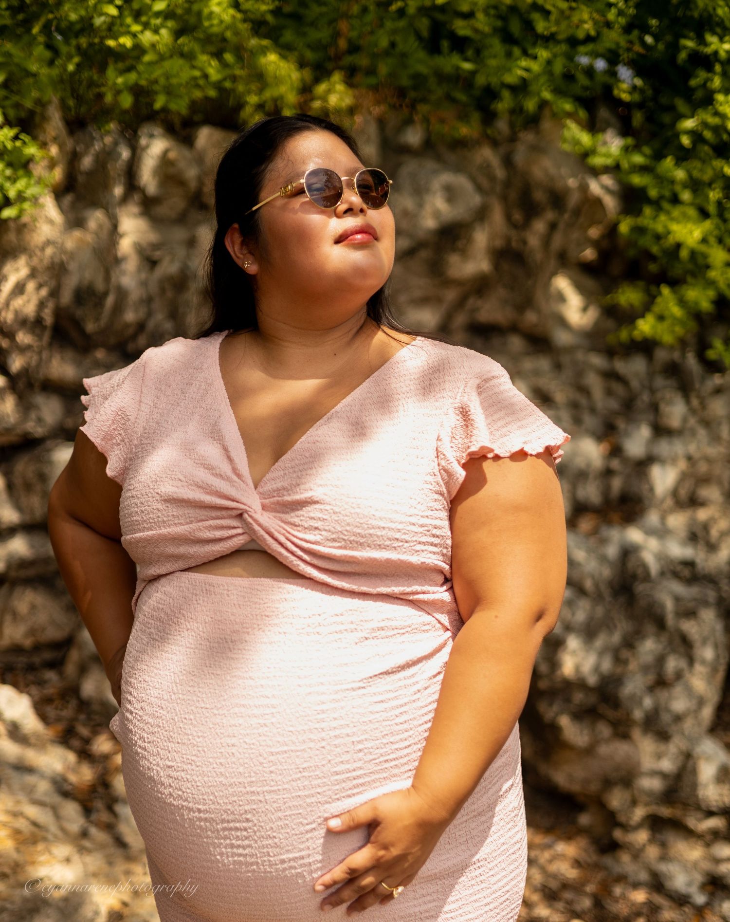 Pregnant woman in pink dress and sunglasses posing outdoors near stone wall on sunny day.