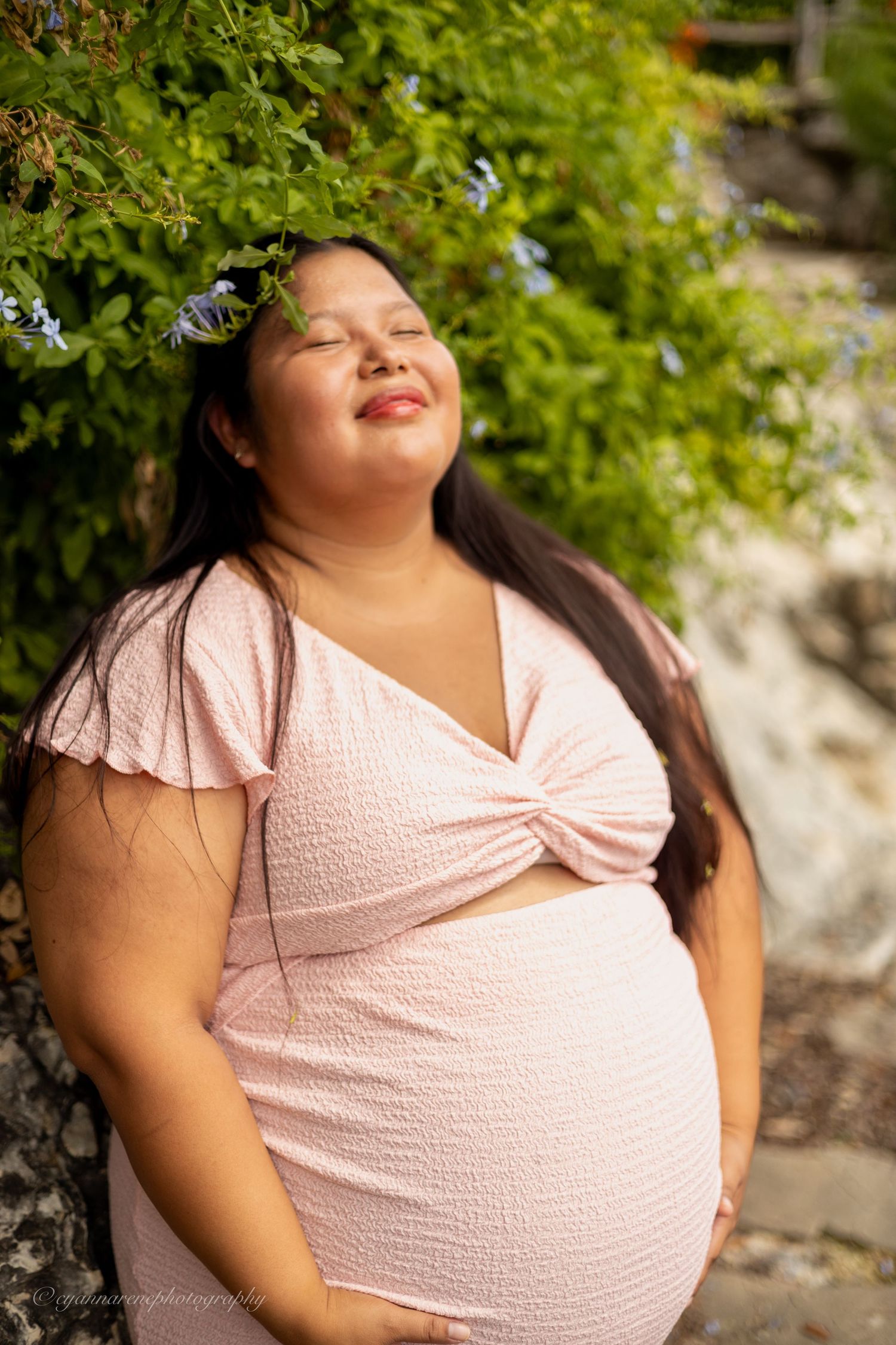 Smiling pregnant woman in pink dress poses outdoors near lush green foliage during a maternity photo shoot.