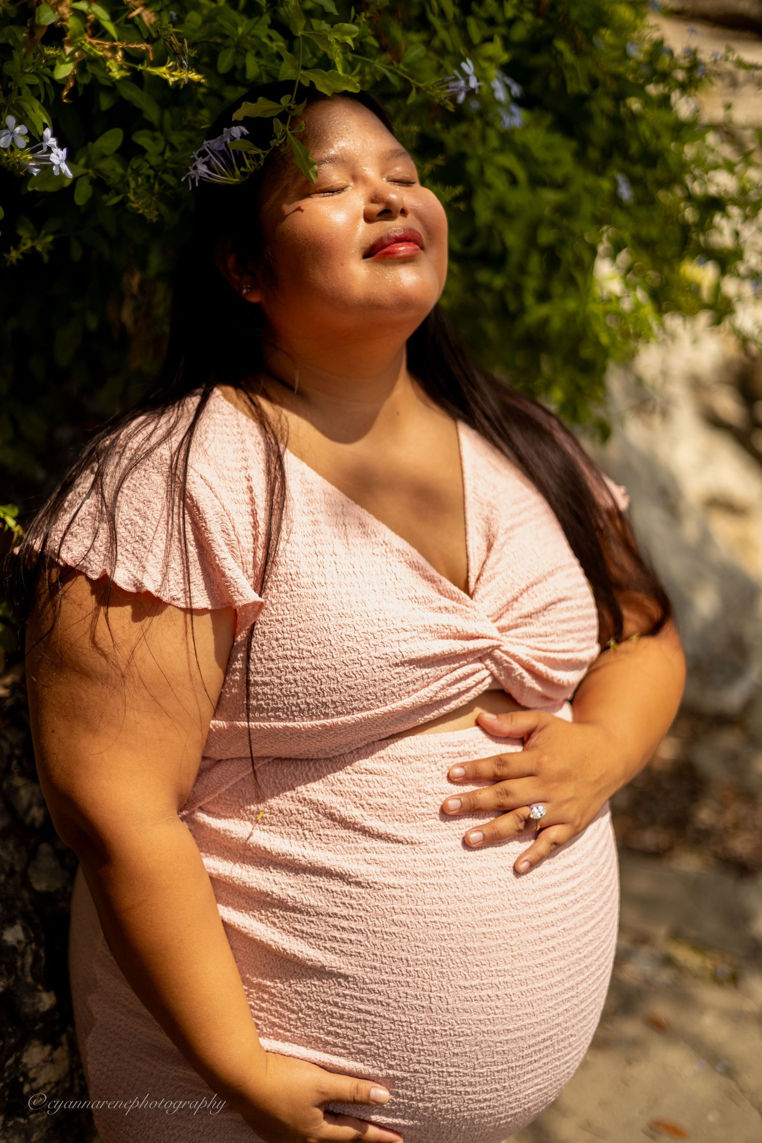 Pregnant woman in pink dress poses outdoors with eyes closed, sunlight glowing behind flowering greenery.