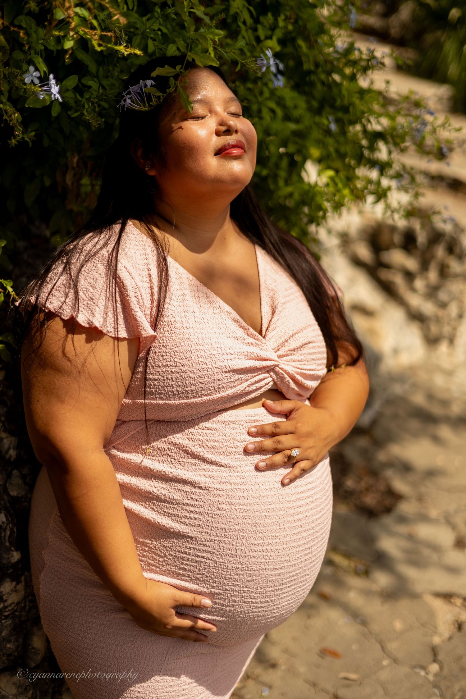 Pregnant woman in pink dress cradling baby bump outdoors near stone wall and lush green foliage in warm sunlight.