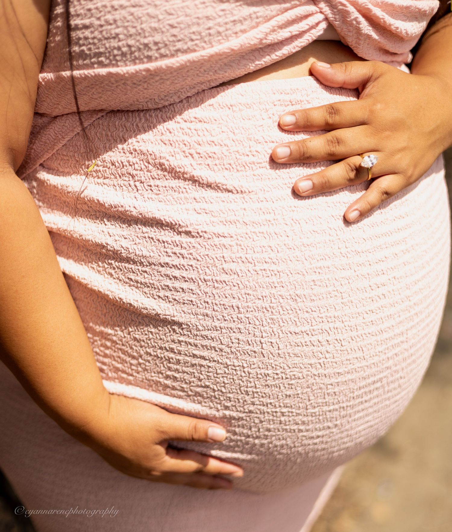 Close-up of a pregnant woman in a pink dress cradling her baby bump, wearing an engagement ring.