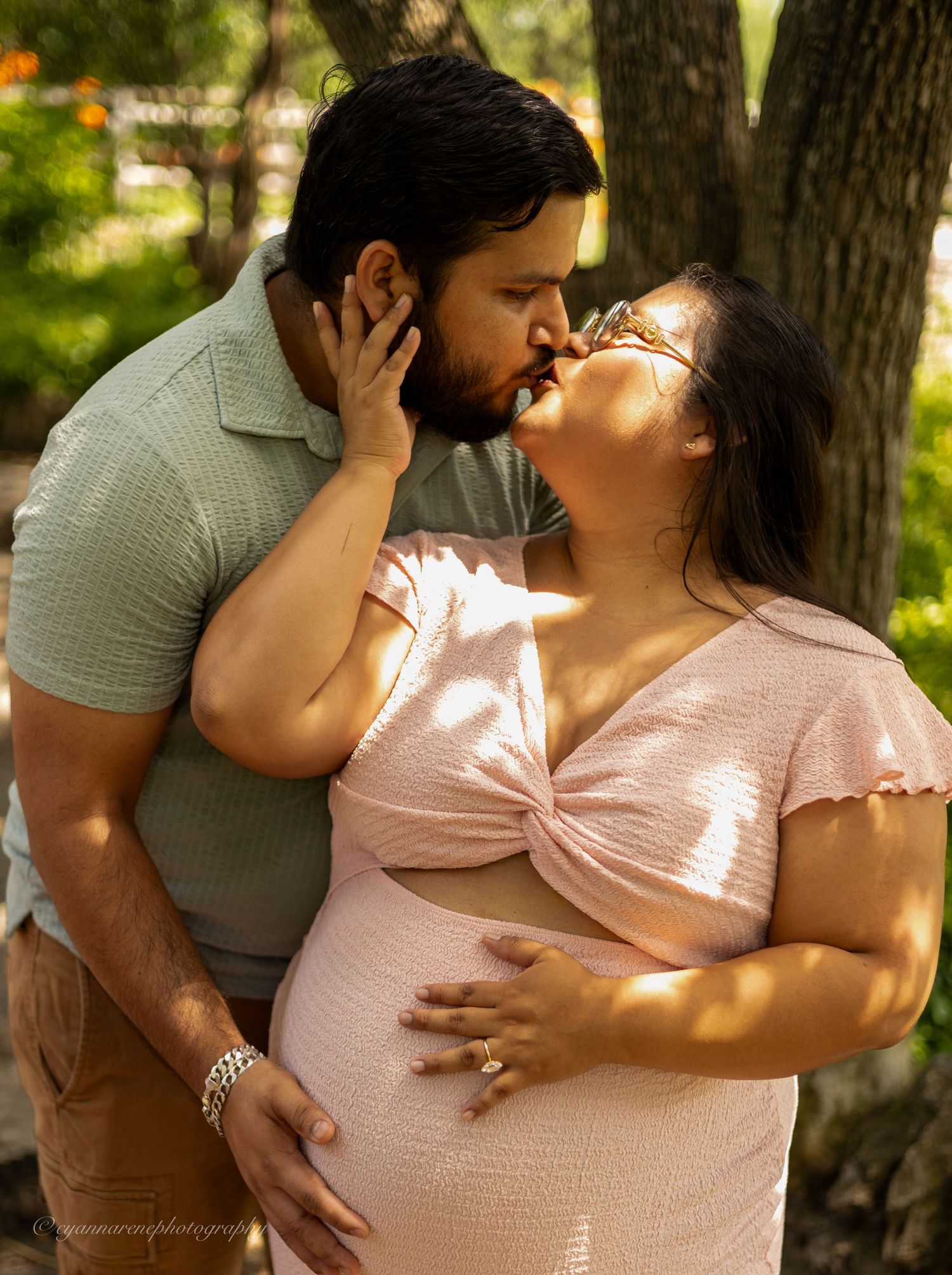 A couple shares a tender kiss outdoors, with the woman visibly pregnant, both dressed casually in a sunlit park setting.