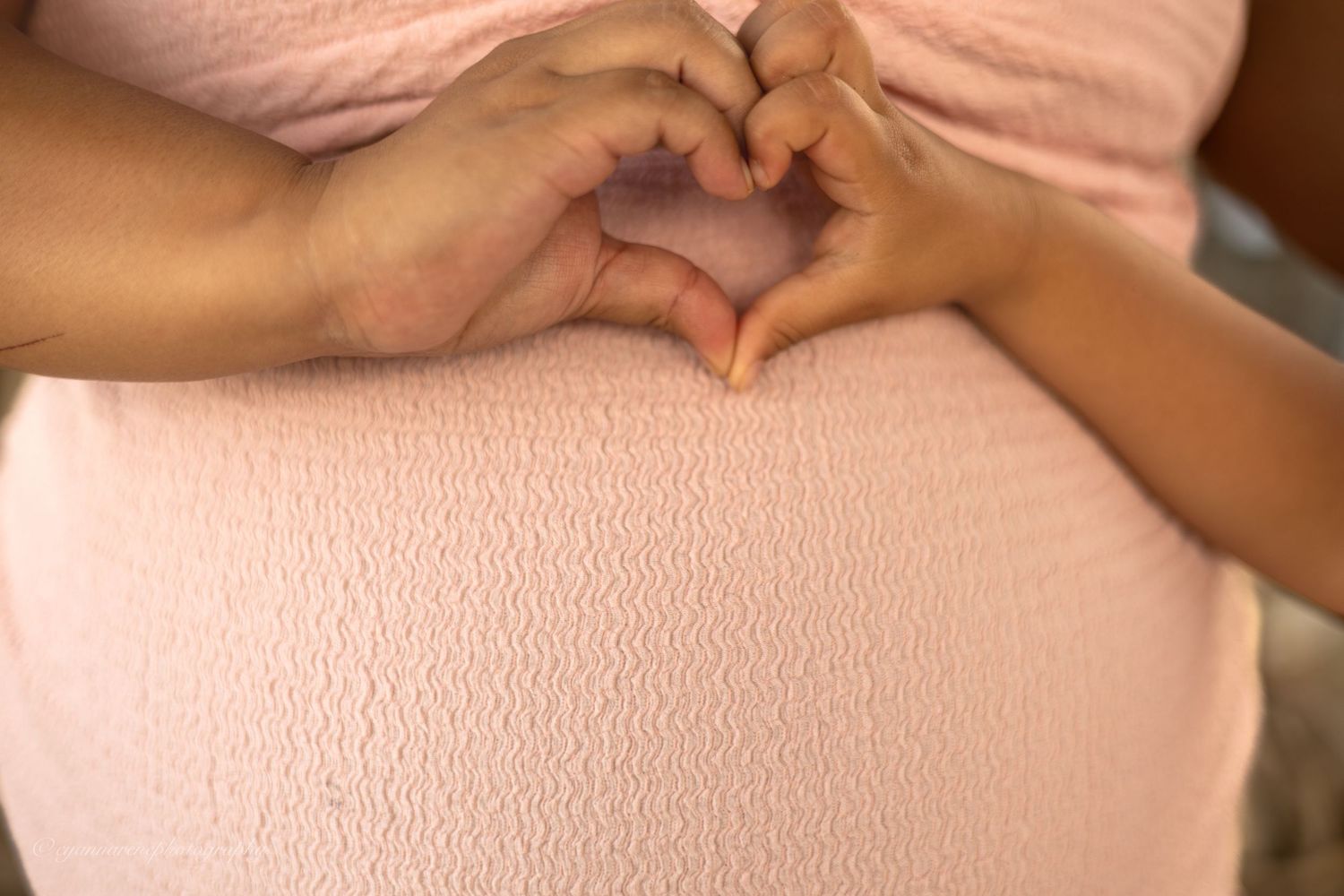 Pregnant woman in pink dress with hands forming a heart shape over her baby bump.