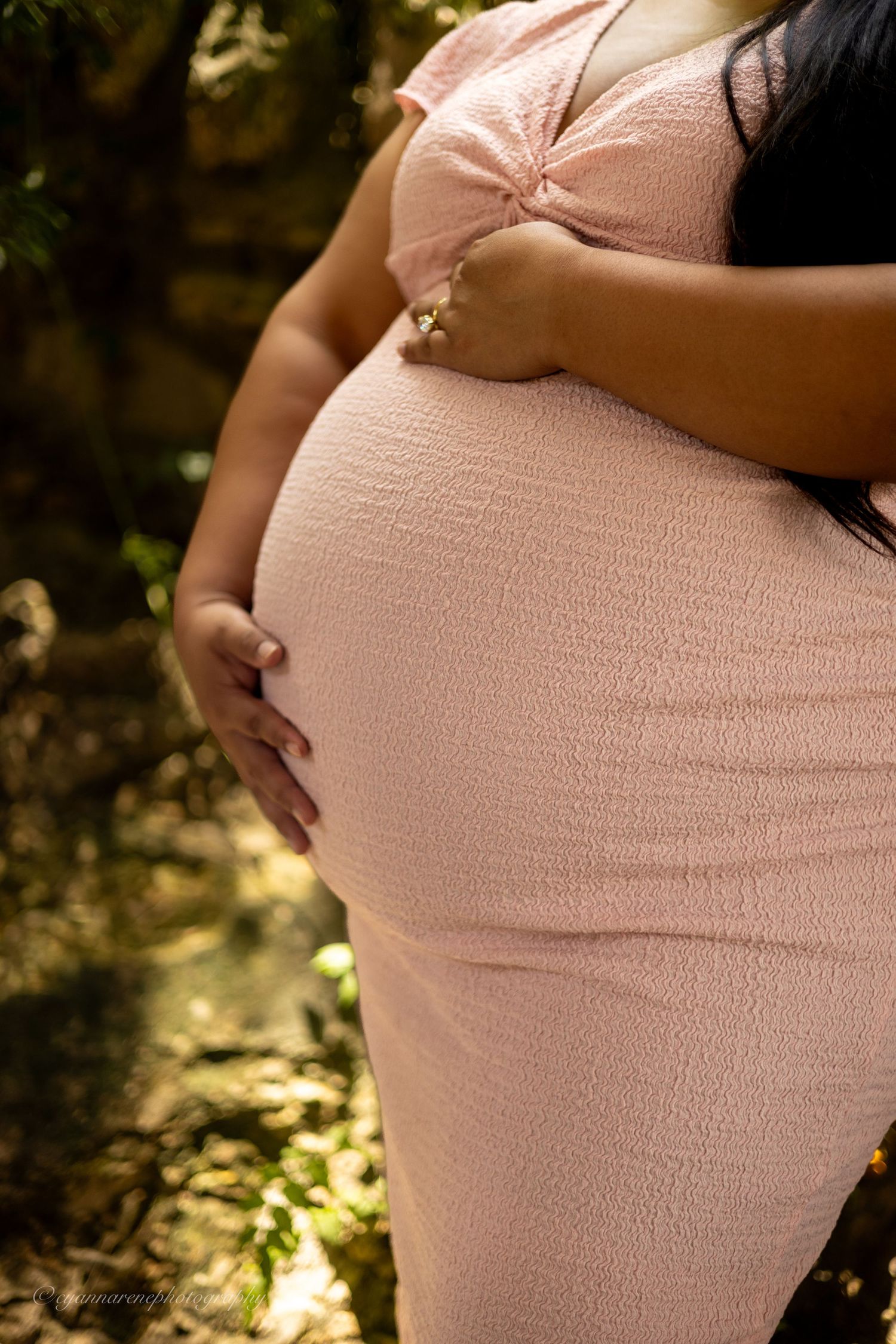 Pregnant woman in pink dress cradling baby bump outdoors in natural woodland setting during maternity photoshoot.