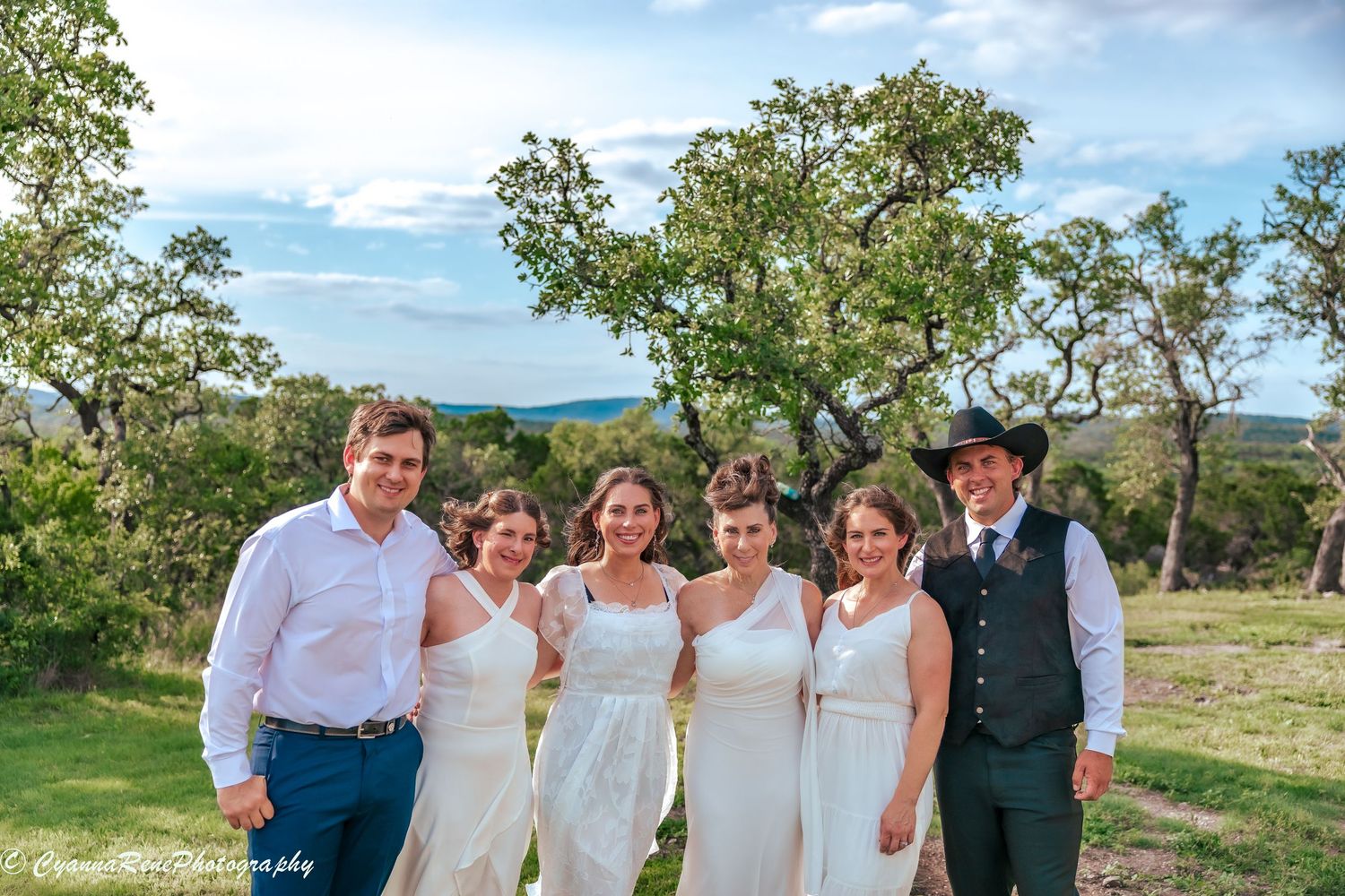 Wedding party poses outdoors with trees and blue sky, featuring brides in white dresses and guests in formal attire.