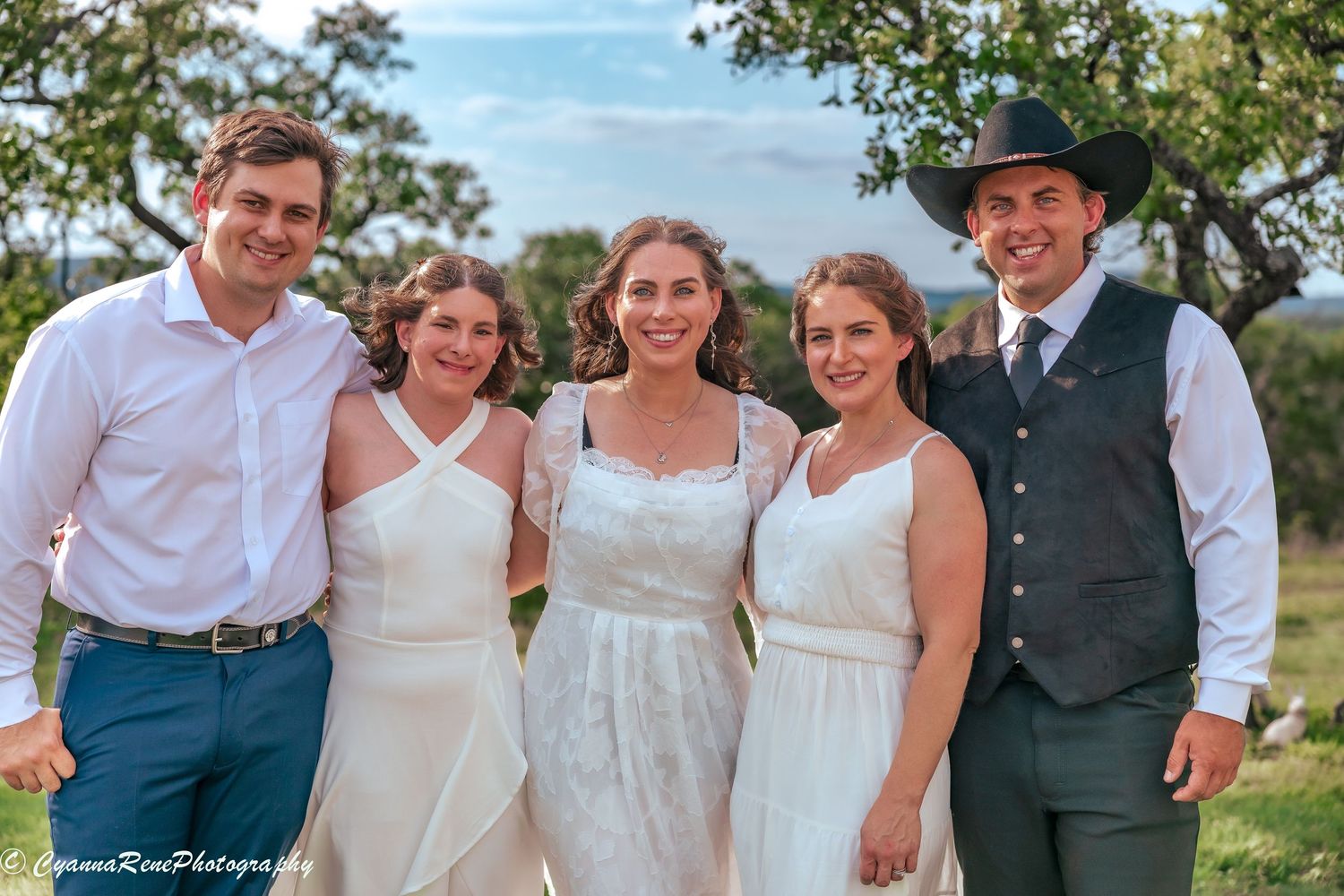 Wedding party of five posing outdoors, three women in white dresses and two men, one wearing a black cowboy hat.