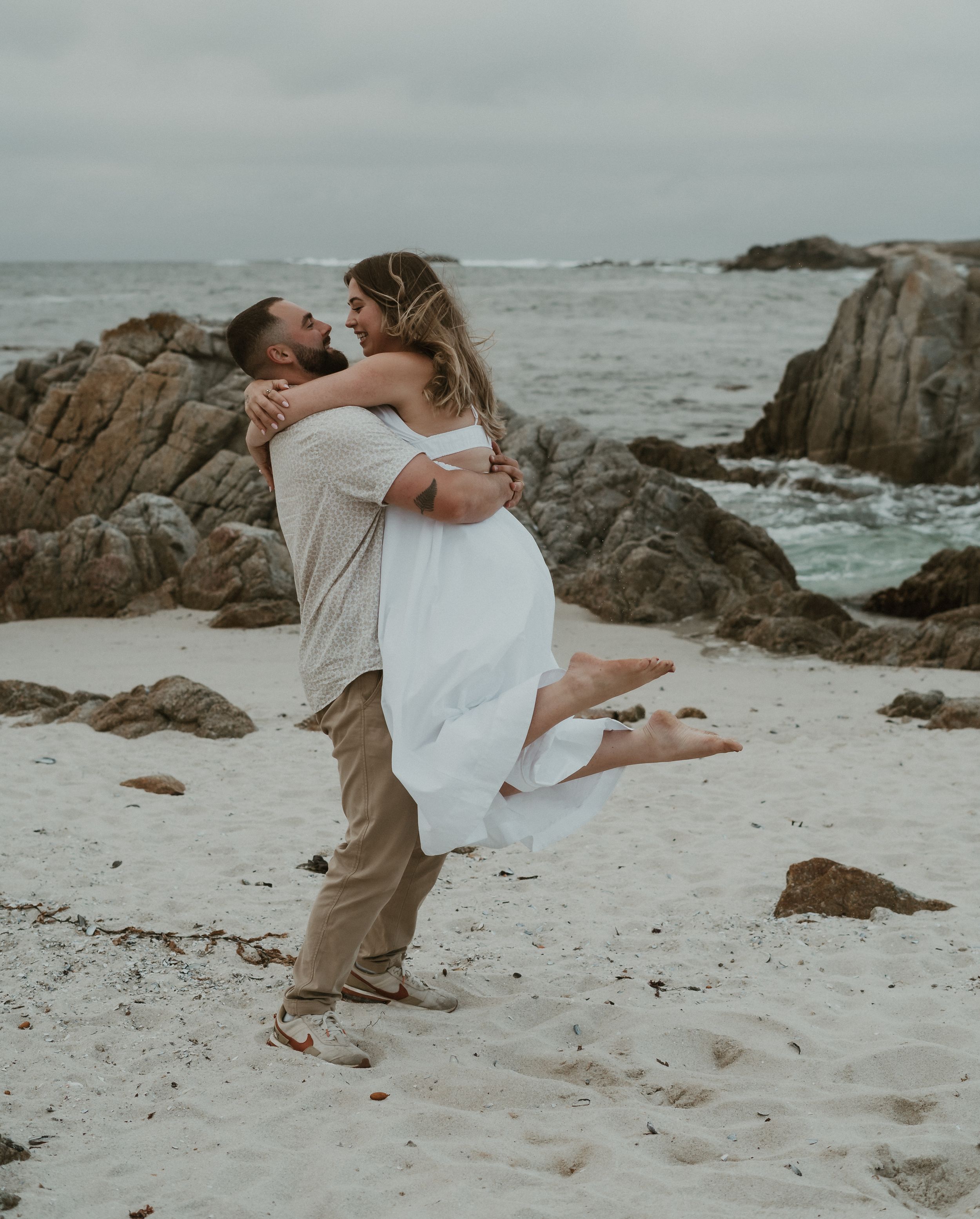 A couple performs dynamic dance moves on a rocky beach with crashing waves and overcast skies in the background.