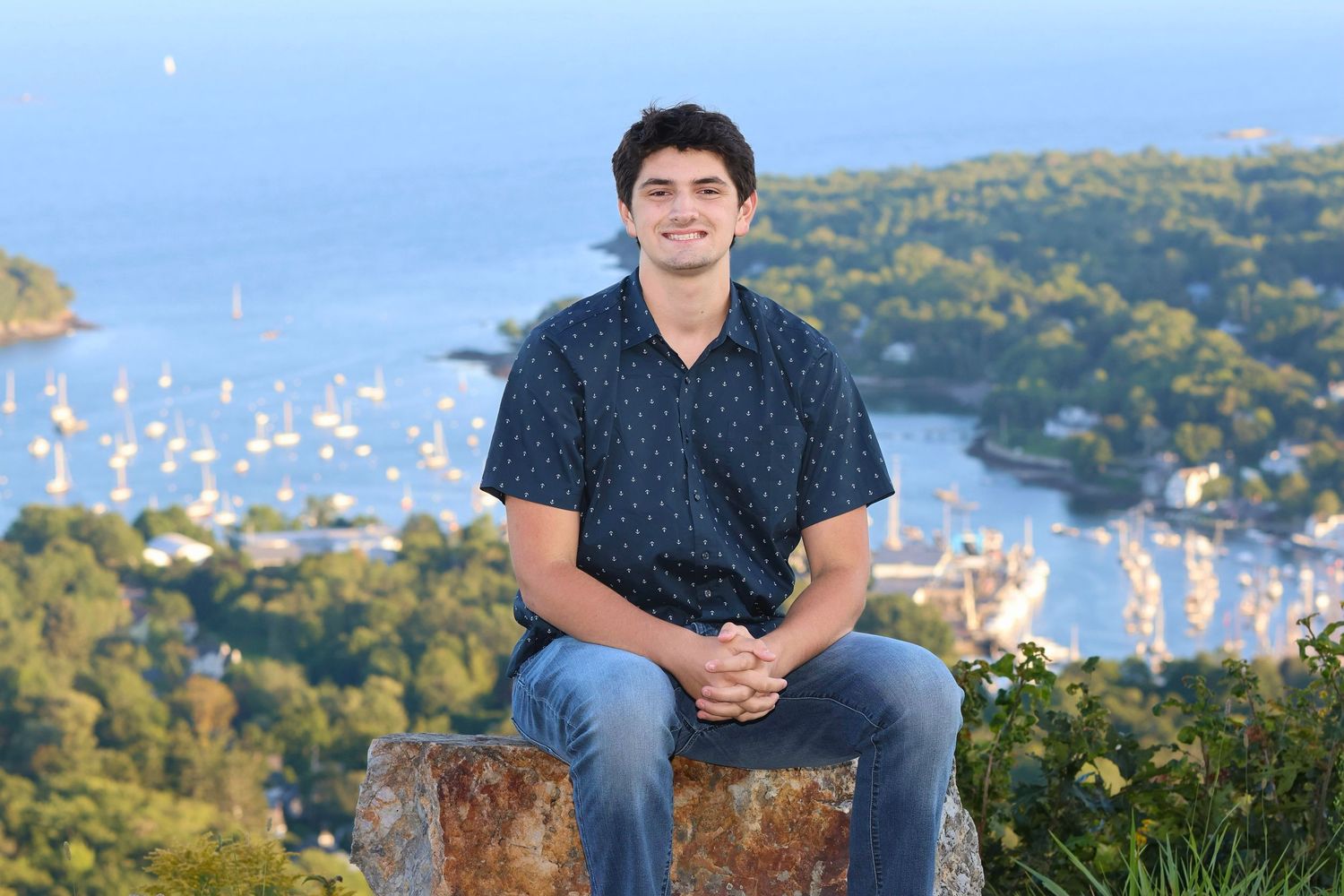 High school senior guy sitting on a rock overlooking a harbor with sail boats in the background wearing jeans and button down short sleeve.