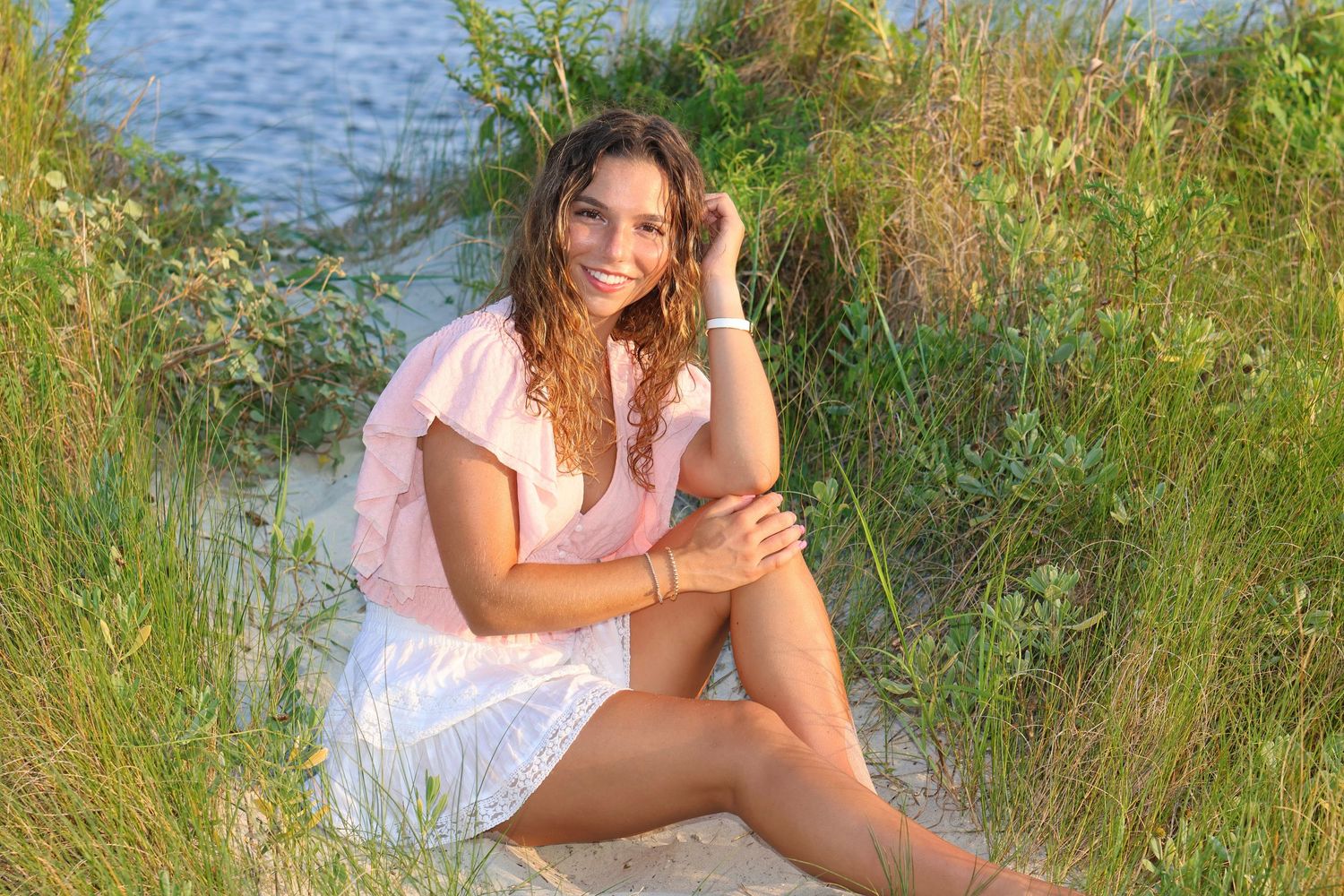 High school senior girl with a short white skirt with pale pink top sitting with leg out in the sand on the beach with water in background.
