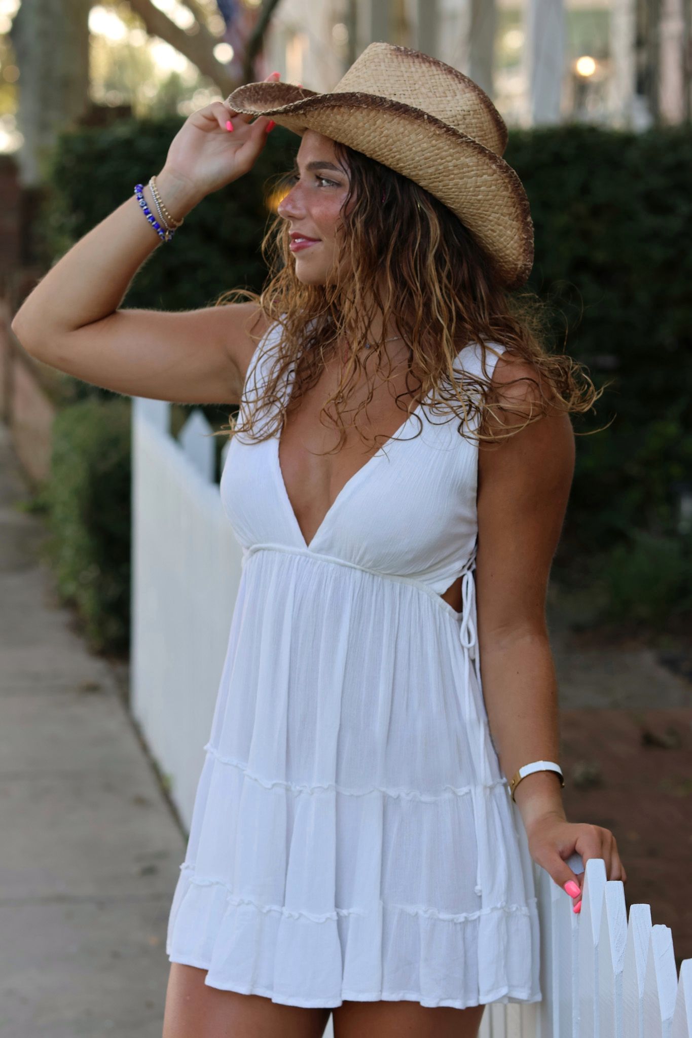 High school senior girl wearing a short white dress and cowboy hat at sunset with a white picket fence behind her.