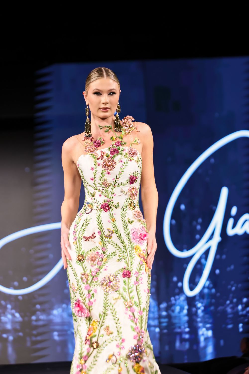 A flowery dress is showcased on the runway against a dark blue backdrop with illuminated signage.