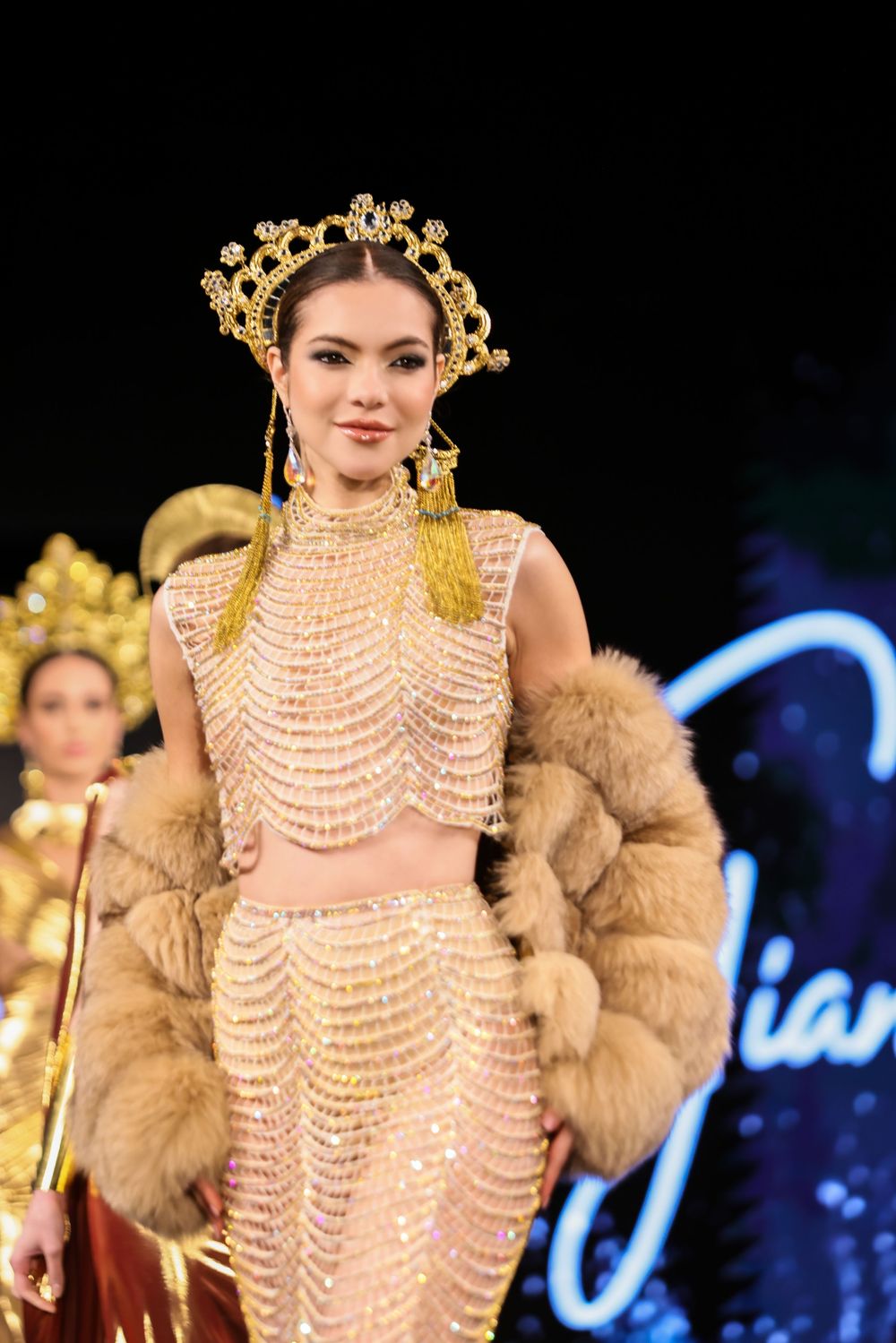Model walks runway wearing layered pearl necklaces and golden crown with tan fur jacket at fashion show.