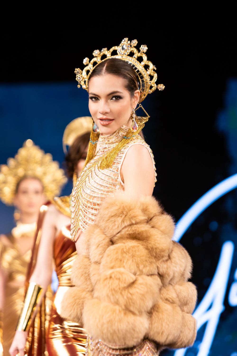 Model walks runway in luxurious fur coat with ornate gold crown and pearl necklaces at fashion show event.