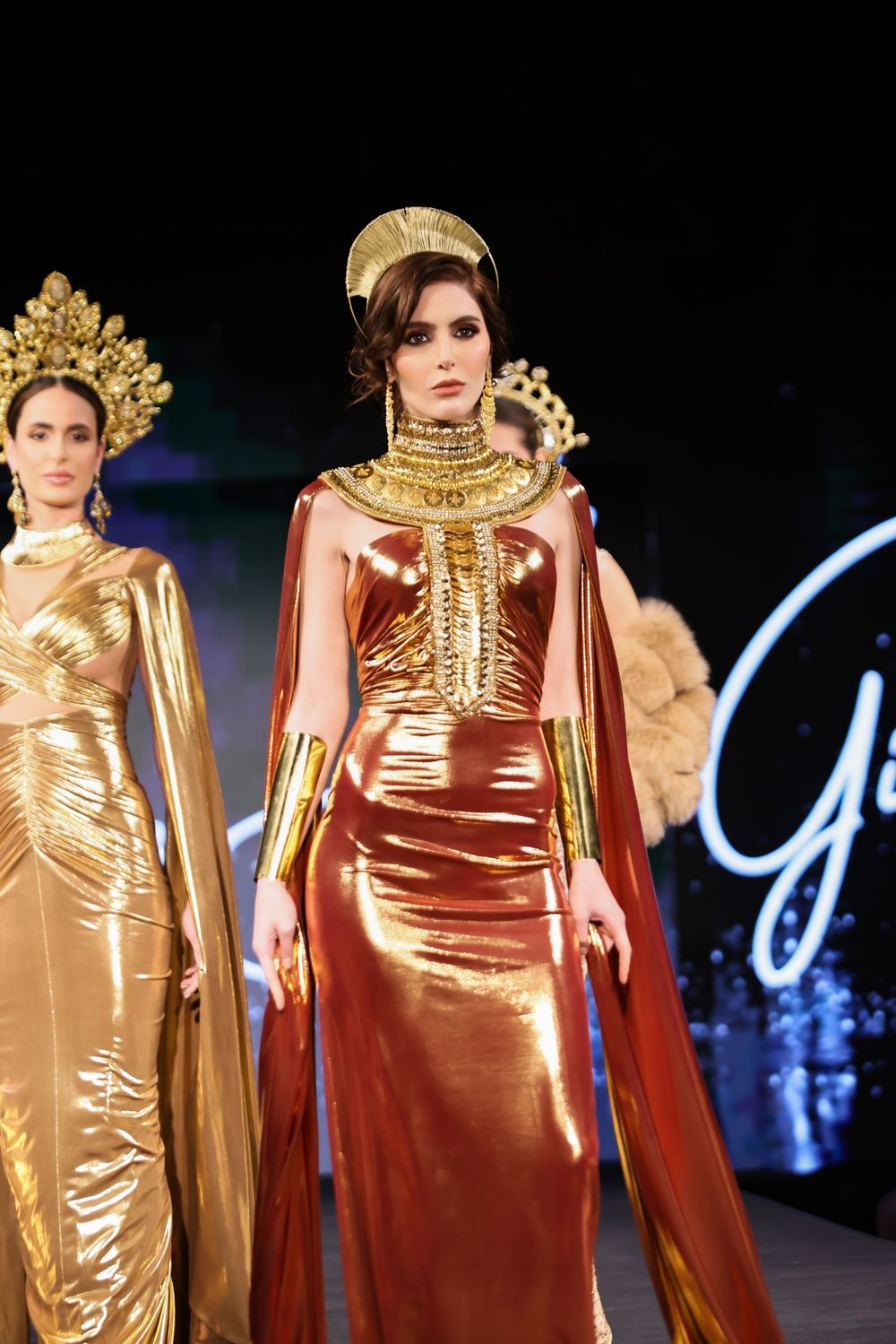 Models walk the runway wearing luxurious golden gowns with flowing capes at a fashion show.