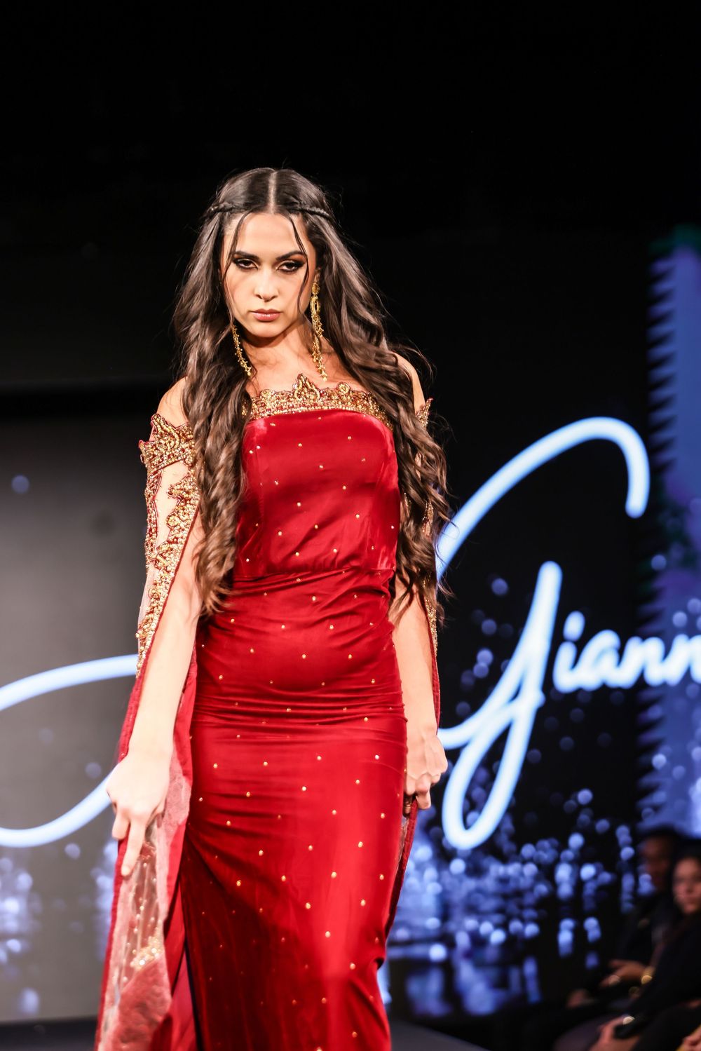 Model showcases a stunning red beaded gown with sheer sleeves during a fashion show on a dark runway.