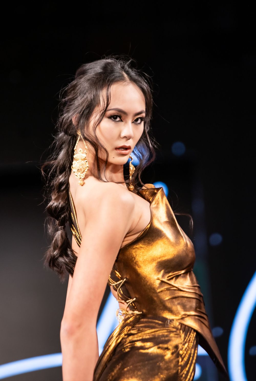 Fashion model walks runway in shimmering gold metallic dress against dark background with blue lighting.