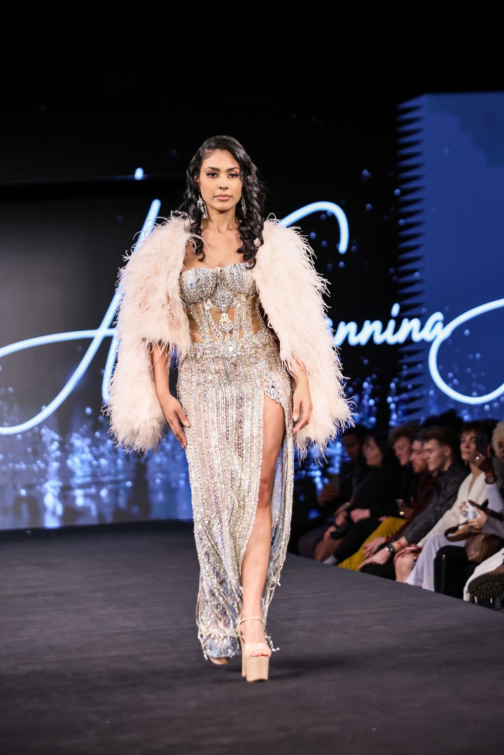 Model walks runway in sparkly silver evening gown with dramatic white feather shoulders at fashion show.