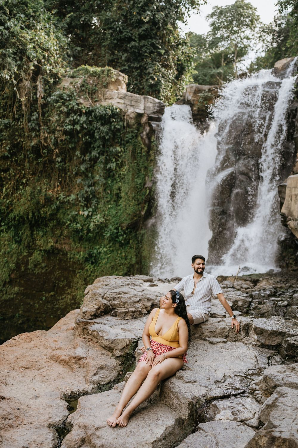 Couple relaxing on rocky outcrop with tropical waterfall view.