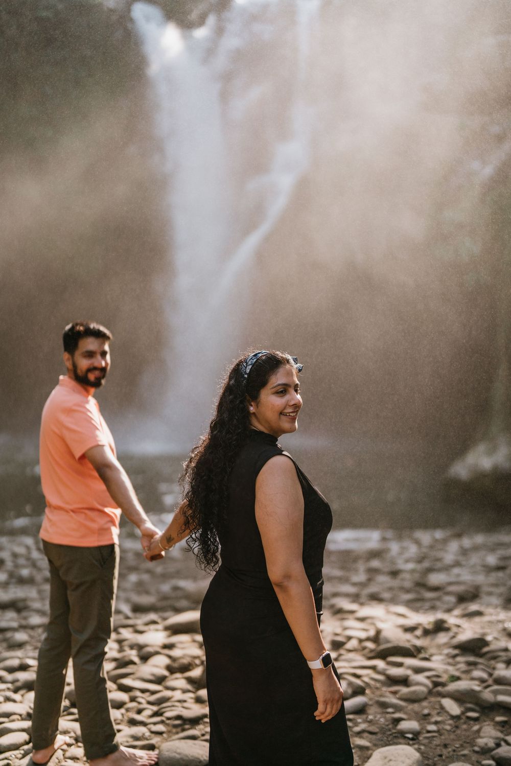 Two people share playful moments on rocky terrain near a cascading waterfall.