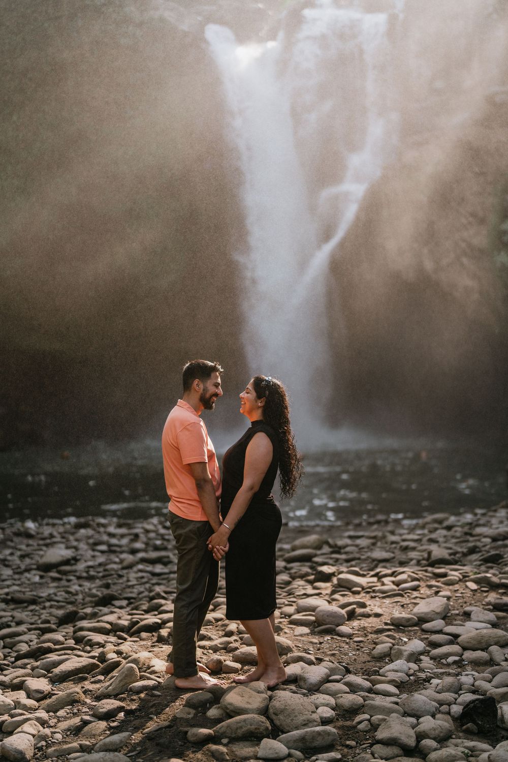 Couple standing on rocky riverbed with misty waterfall cascading in background.