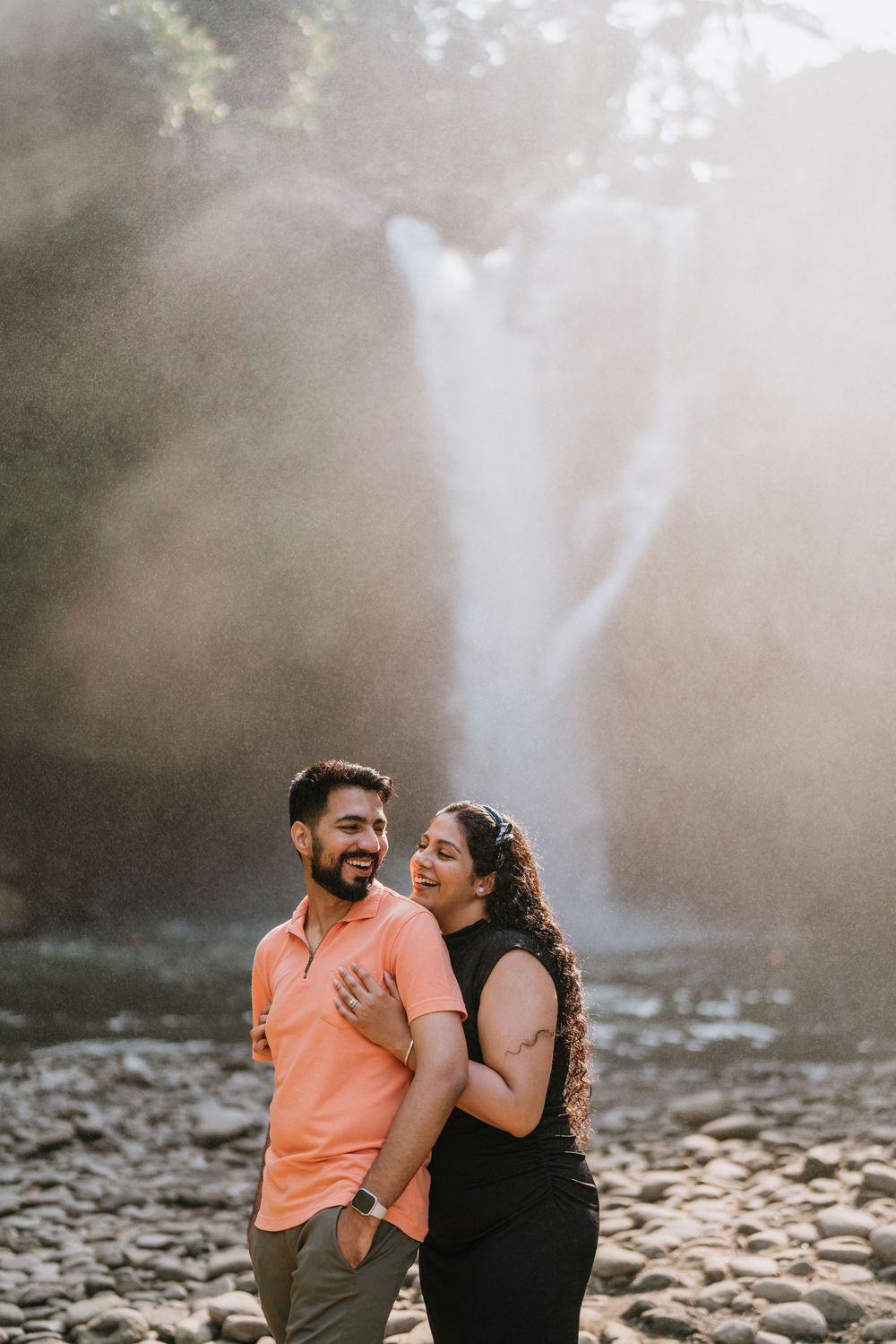 Couple laughing together on rocky shore with dramatic waterfall and sunlight in background.