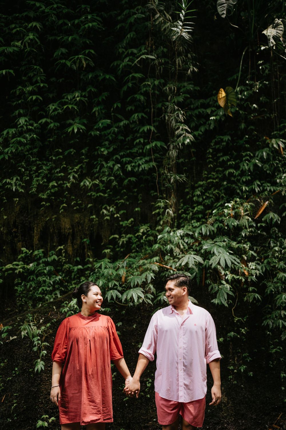 Two people in coral and pink outfits walking through a lush green forest setting.