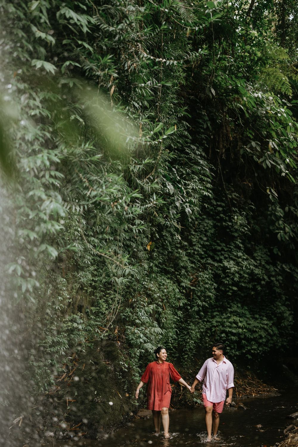 A couple walks hand in hand through a lush green forest path.