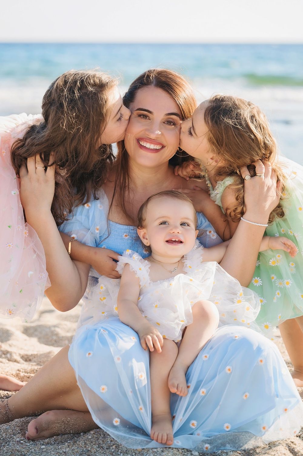 Family enjoying tender moments together while sitting on a sunny beach wearing light blue and white outfits.