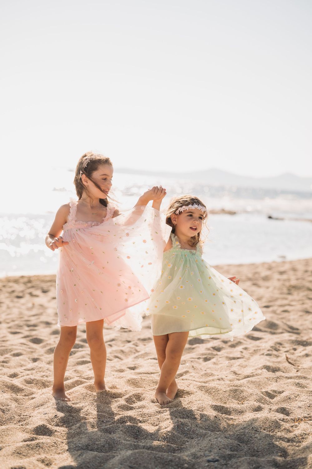 Two small children in flowy dresses dance and play together on a sunny beach.