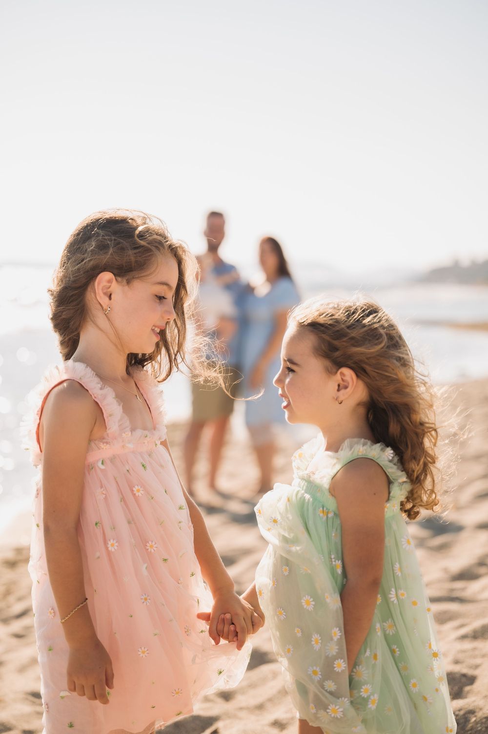 Two young children in pastel dresses share a sweet moment together on a sunny beach.