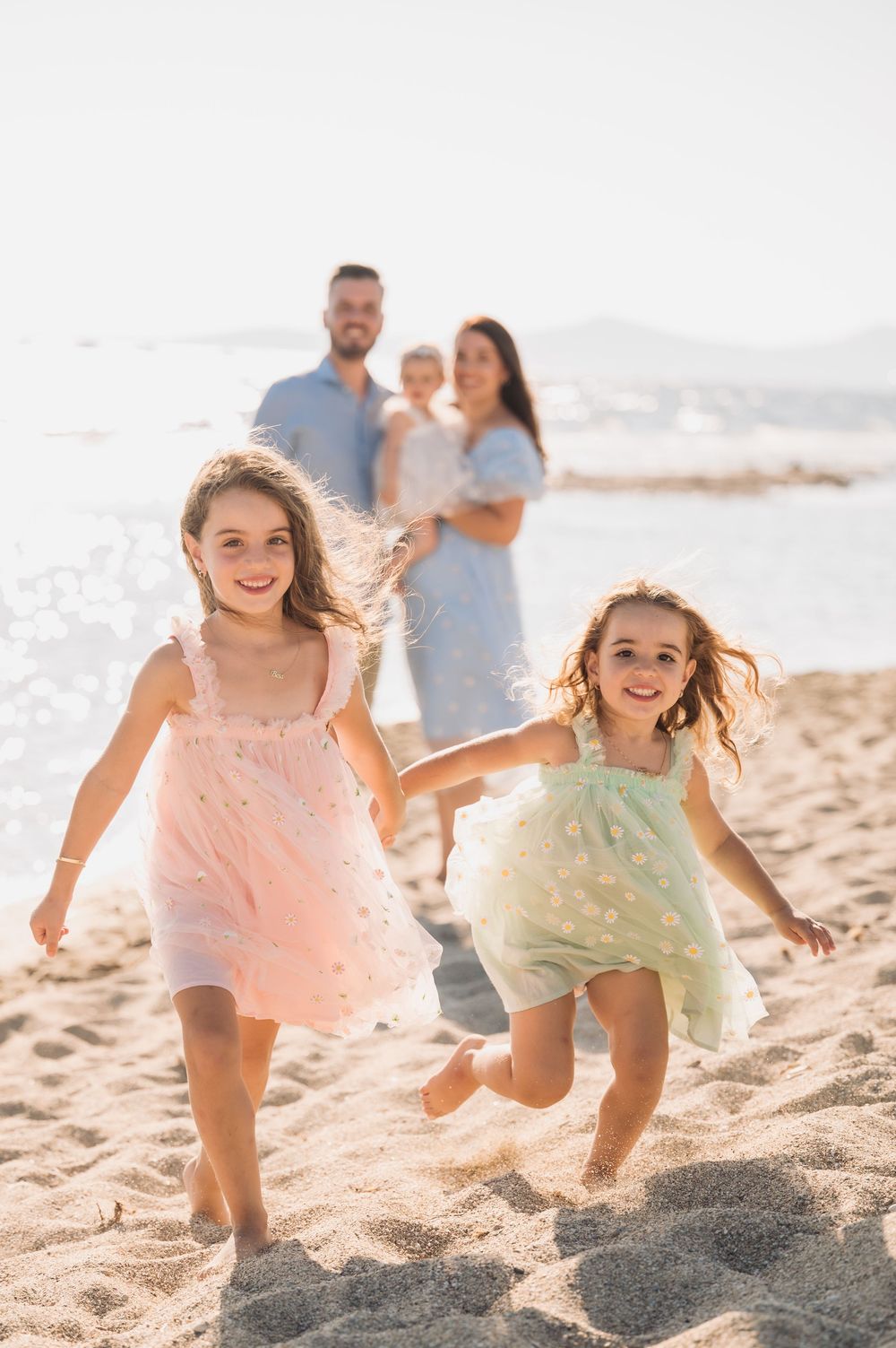 Two children in pastel dresses run and jump playfully along a sunny beach with family in background.