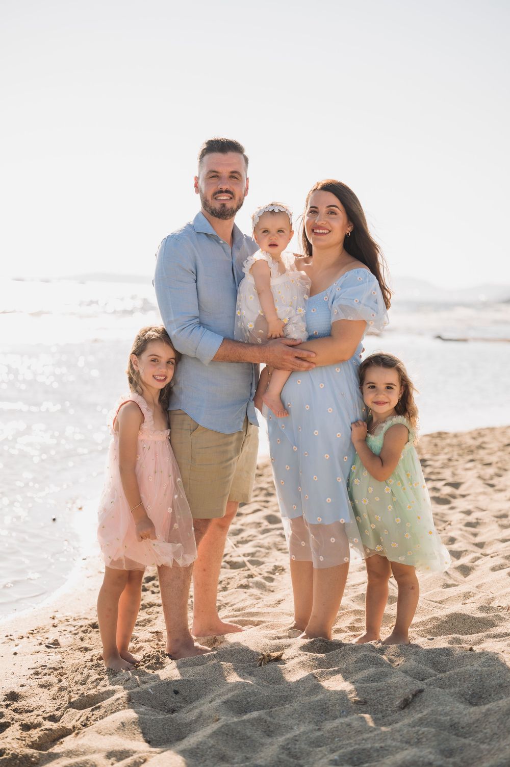 A family stands together on a sunny beach wearing light colored clothing and enjoying the seaside atmosphere.
