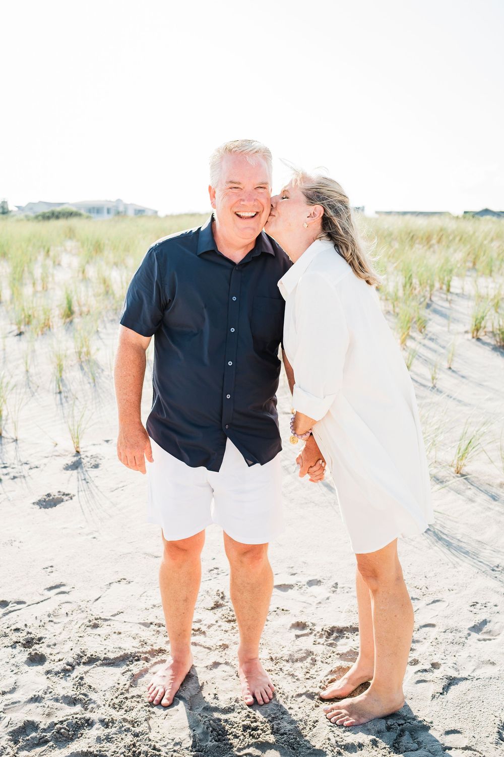 Couple shares intimate moment together on sunny beach with sand dunes in background.
