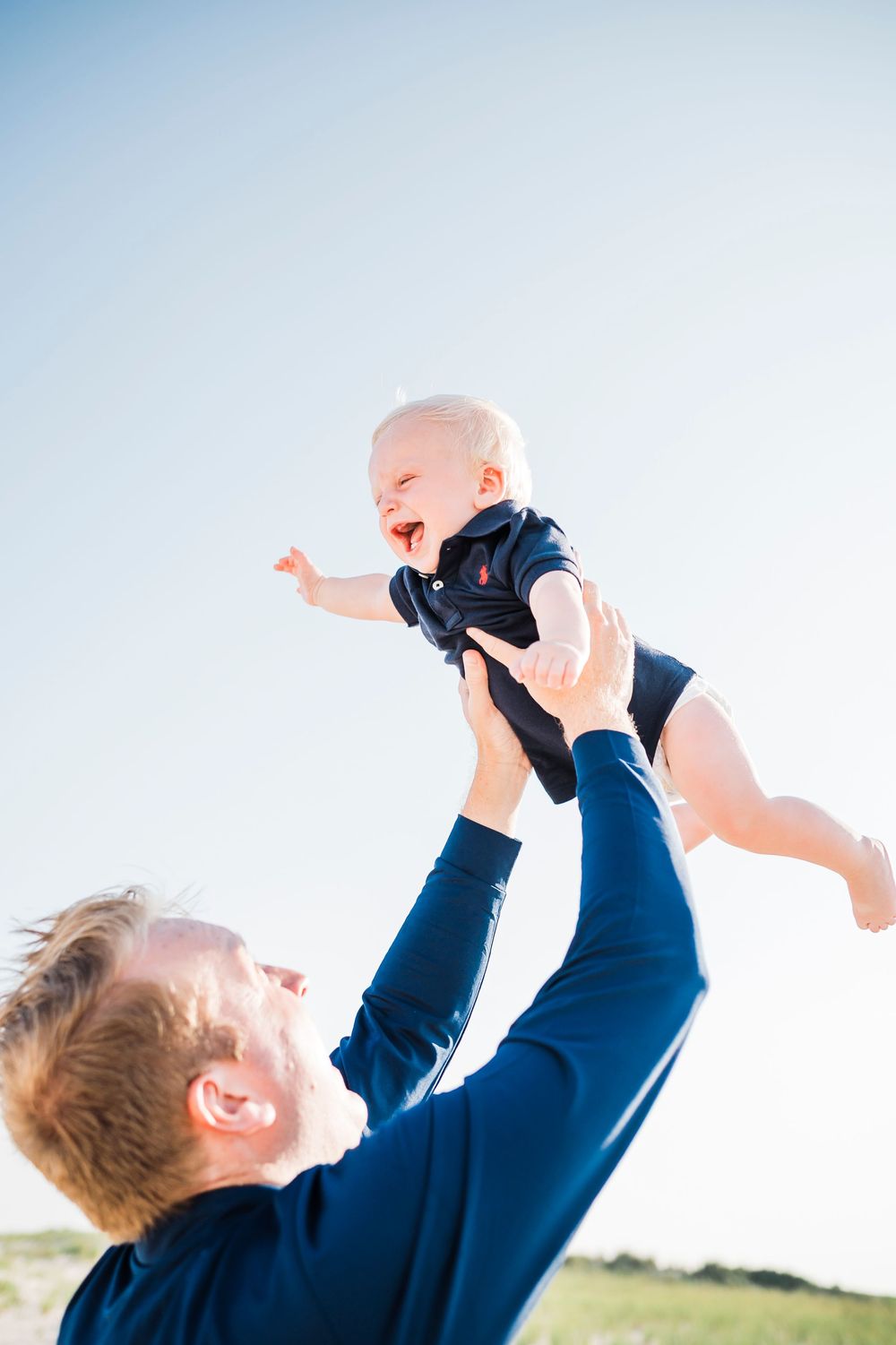 Parent lifts joyful baby wearing navy onesie into blue sky during outdoor playtime.