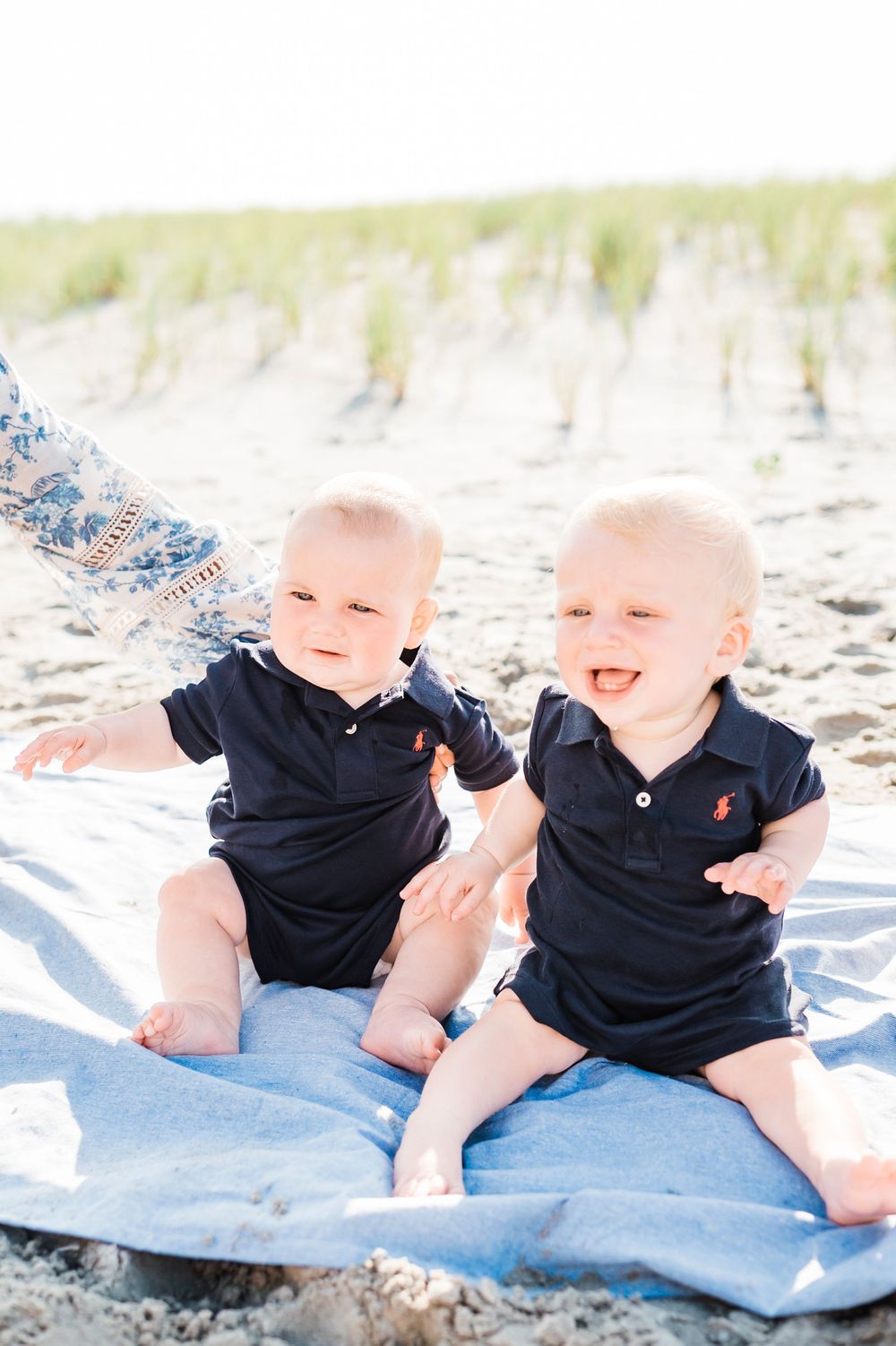 Two babies in matching navy polo outfits sit together on blue blanket at sunny beach.