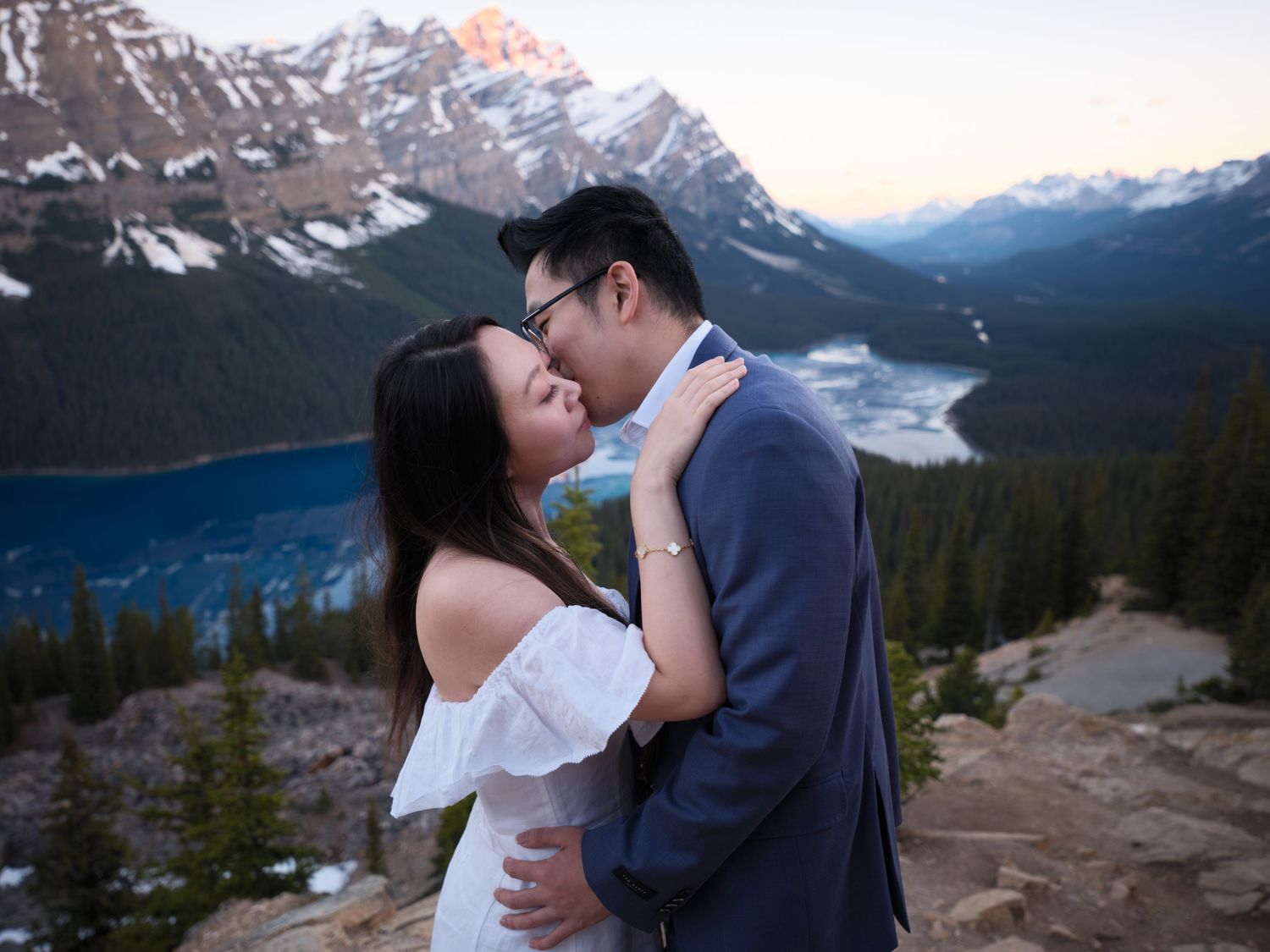 Two figures embrace while standing on a mountain overlook with snowy peaks and glaciers in the scenic backdrop during a spring wedding Banff