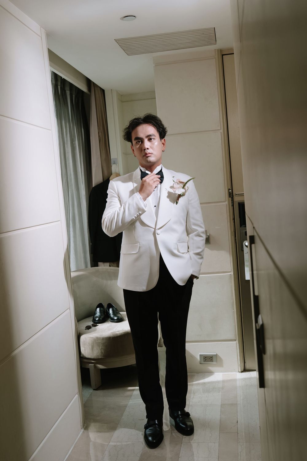 Groom in white tuxedo jacket adjusting bow tie while standing in hotel room mirror.