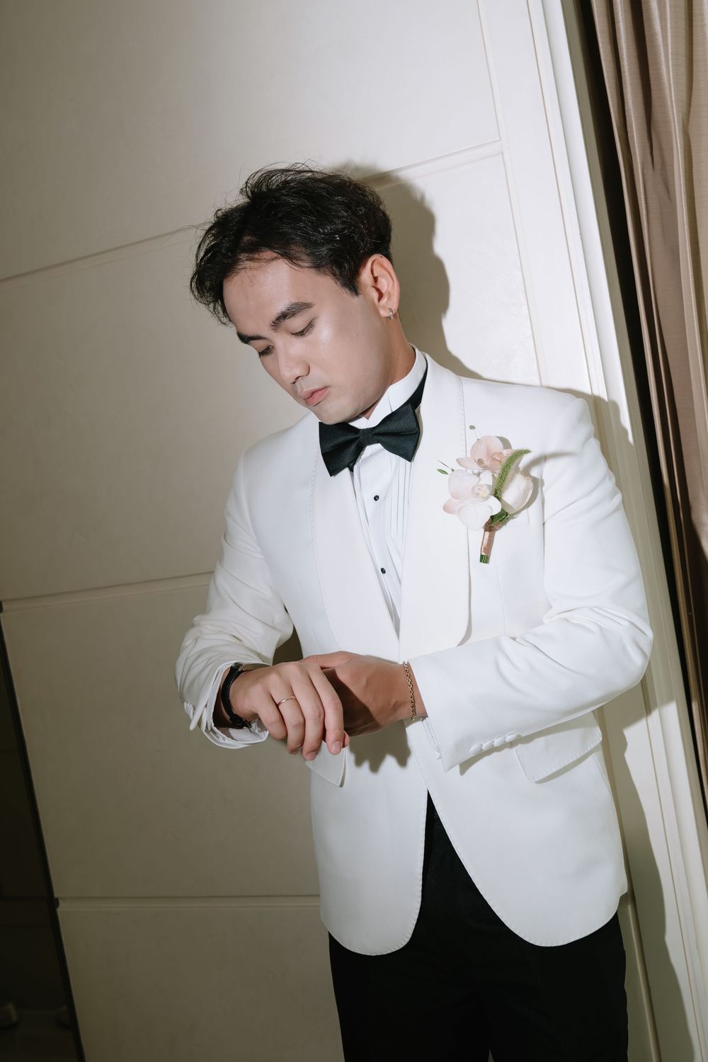 Groom in white tuxedo with floral boutonniere adjusting cufflinks by window before wedding.