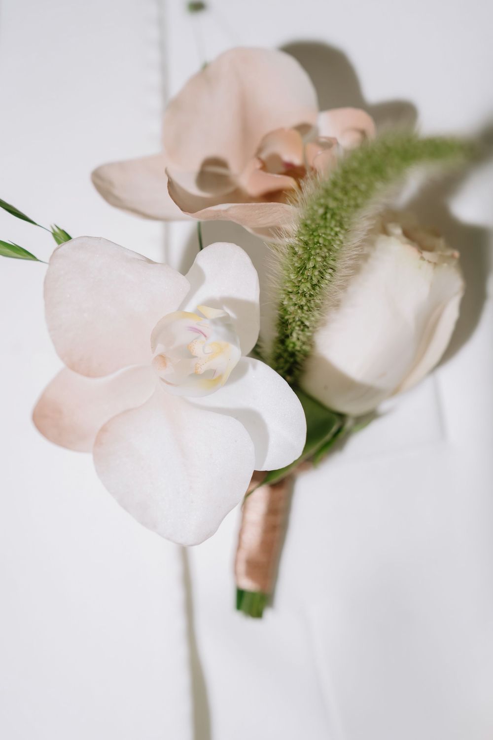 Elegant wedding boutonniere with blush orchid, white tulip, and green accents on white background.