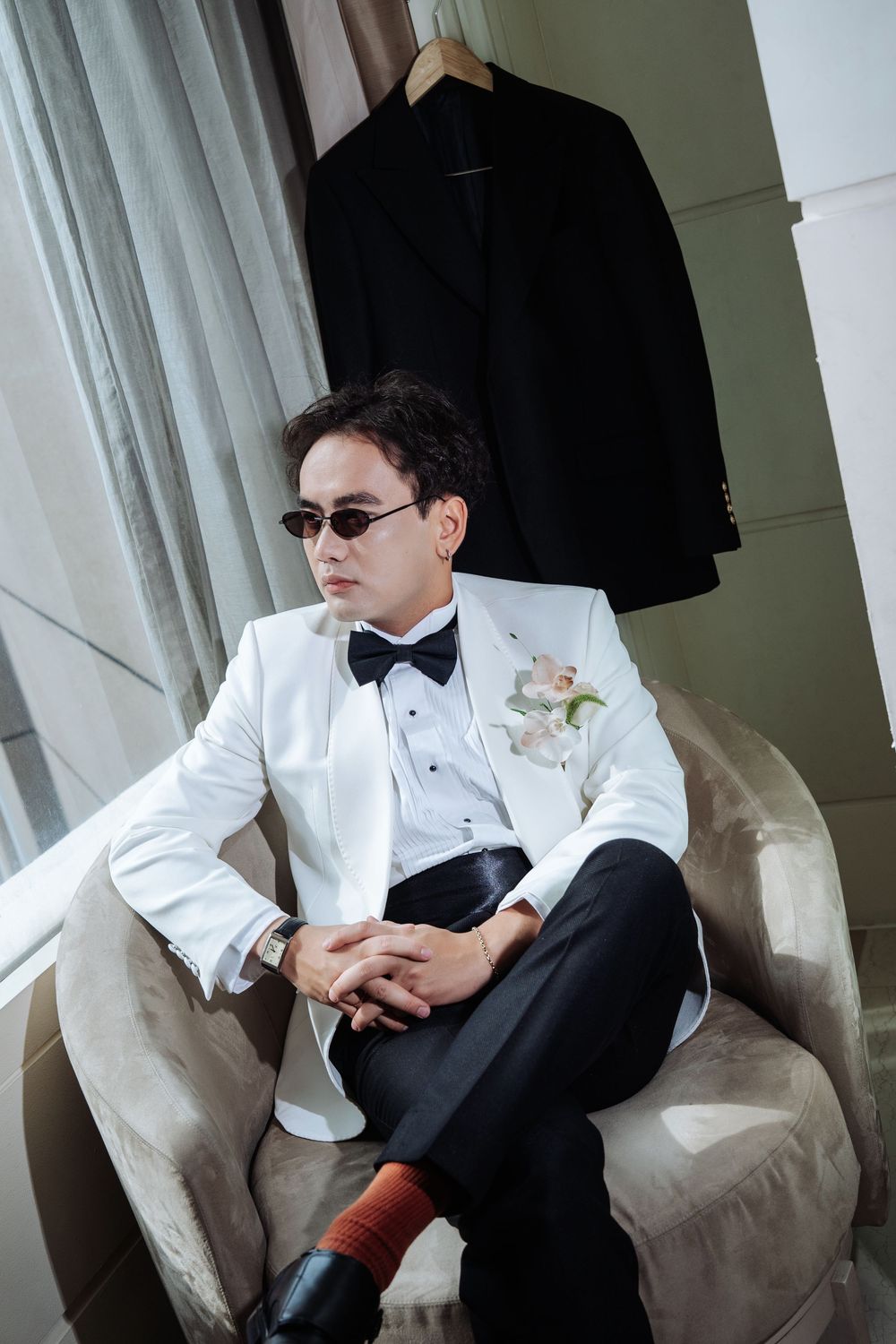 Stylish groom in white tuxedo and sunglasses seated in chair with black suit hanging behind.