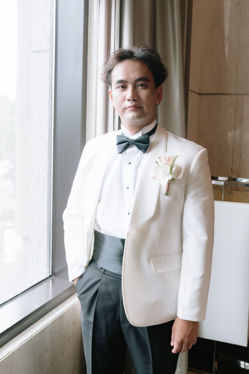 Groom in white tuxedo jacket with boutonniere standing by large window in elegant venue.