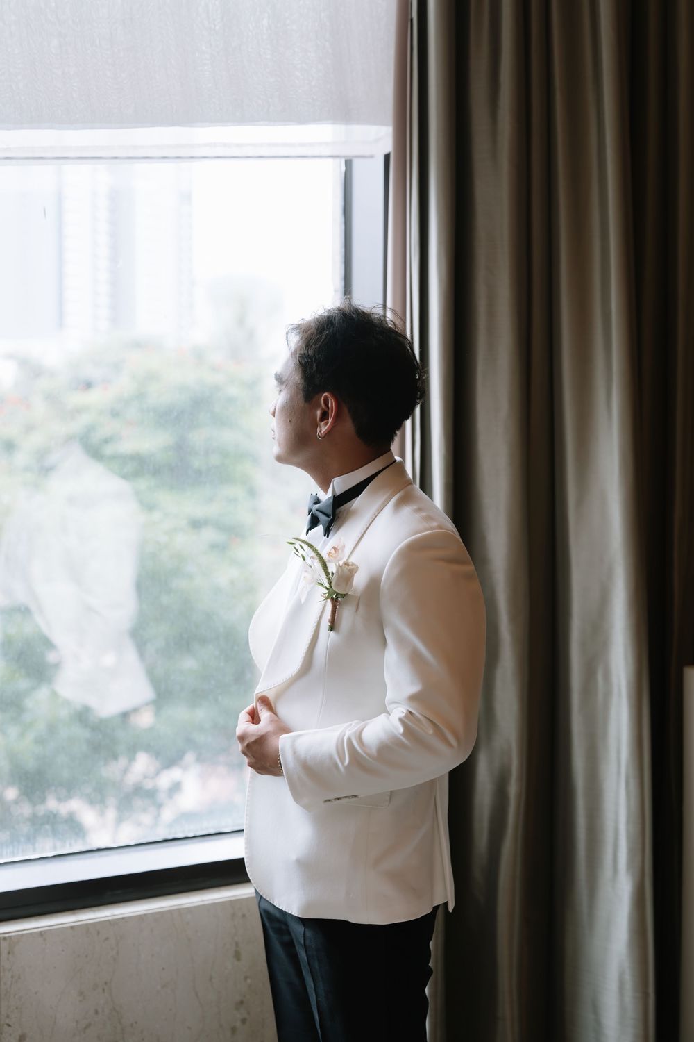 Groom in white tuxedo gazing out hotel window while adjusting jacket before wedding ceremony.