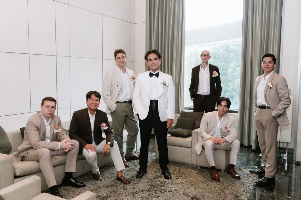 Groom in white tuxedo stands confidently with groomsmen in beige and brown suits in hotel suite.