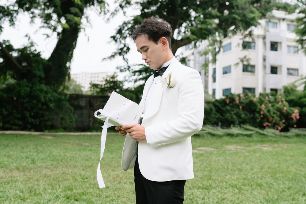 Groom in white tuxedo reading wedding vows from a ribbon-tied booklet outdoors on green lawn.