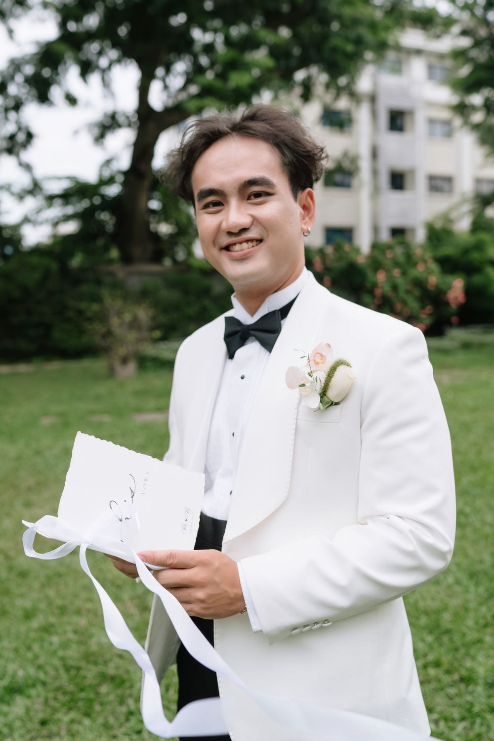 Smiling groom in white tuxedo with boutonniere holding ribbon-tied vow booklet outdoors.